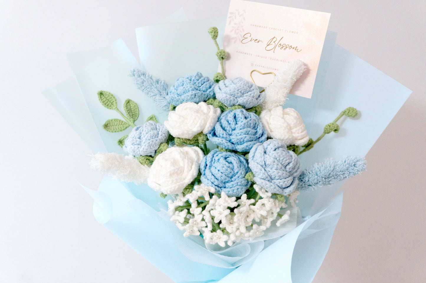這是一束精緻的手工編織玫瑰滿天星花束,採用粉藍、粉紅、紅色、紫色、橙色五種顏色可選,展現繽紛多彩的視覺效果。花束以藍色薄膜包裝,搭配白色和綠色葉片裝飾,中心點綴可愛的蝴蝶結,附帶「Ever Blossom」品牌標籤,極具浪漫氣息。適合用作情人節、生日等特殊場合的禮物,設計細膩,充滿溫暖與創意,令人心動。