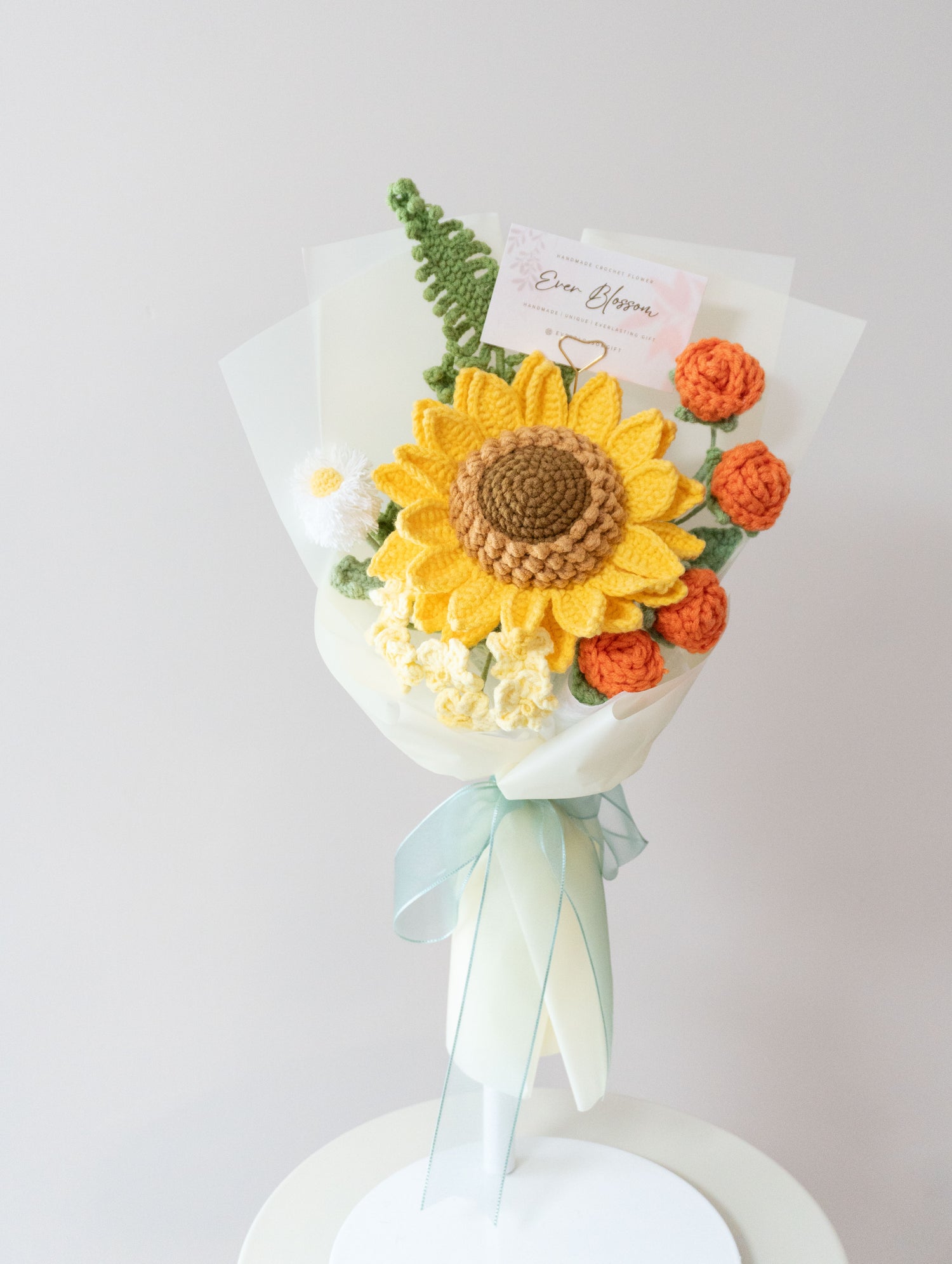 這是一束由香港花店Ever Blossom精心製作的手工編織永生花束，包含鮮艷的向日葵和橙色玫瑰，搭配白色雛菊與綠葉，包裹在淡雅薄紙和藍色緞帶中。花束設計精緻，適合用作謝師宴或感謝長輩的禮物，展現溫暖與感恩的情感。永生花採用高品質材料製作，保存時間長，可作為紀念謝師之情的完美選擇，體現對老師或長輩的尊重與心意。