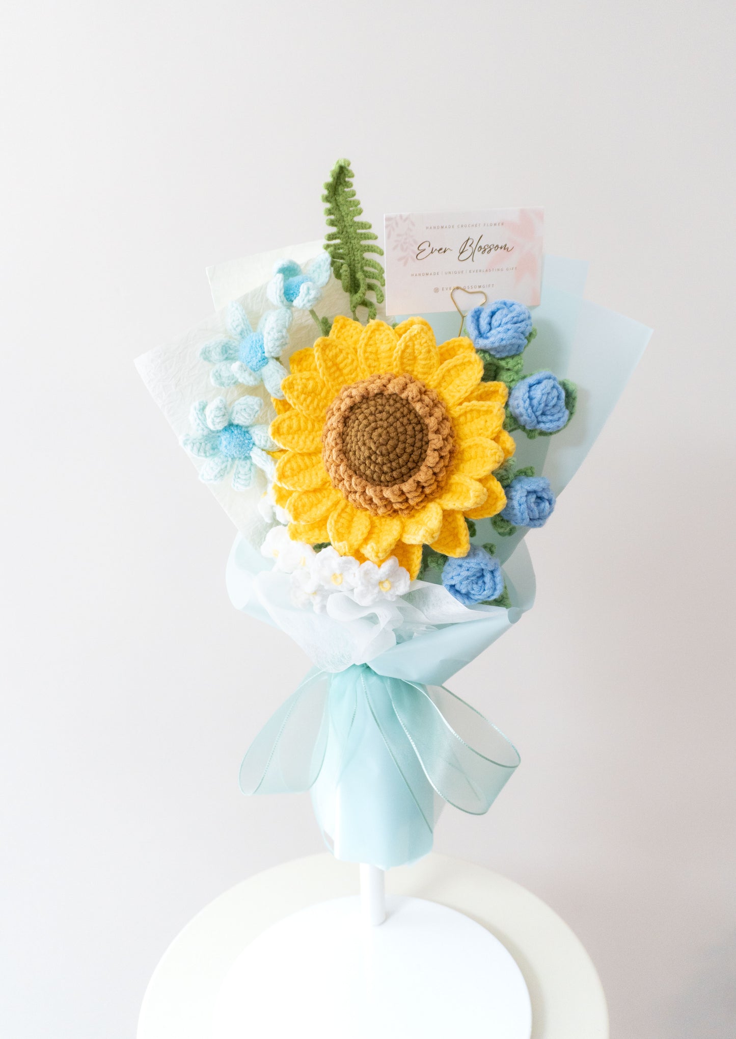 這是一束精心製作的永生花束，由香港花店Ever Blossom提供，結合了鮮艷的向日葵和藍色玫瑰，搭配白色小花和綠葉，包裹在淡雅的薄紙和藍色緞帶中。花束設計精緻，適合用作生日、紀念日或表達愛意的送禮選擇，展現溫暖與浪漫的情感。這款永生花採用高品質材料製作，保存時間長，可作為家居裝飾或送給親友的完美禮物，體現香港花店的專業工藝與用心。
