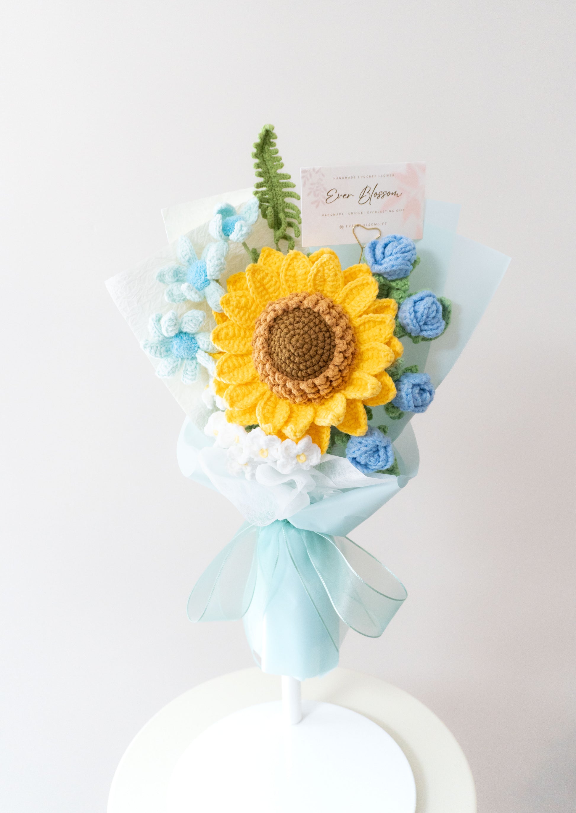 這是一束精心製作的永生花束，由香港花店Ever Blossom提供，結合了鮮艷的向日葵和藍色玫瑰，搭配白色小花和綠葉，包裹在淡雅的薄紙和藍色緞帶中。花束設計精緻，適合用作生日、紀念日或表達愛意的送禮選擇，展現溫暖與浪漫的情感。這款永生花採用高品質材料製作，保存時間長，可作為家居裝飾或送給親友的完美禮物，體現香港花店的專業工藝與用心。
