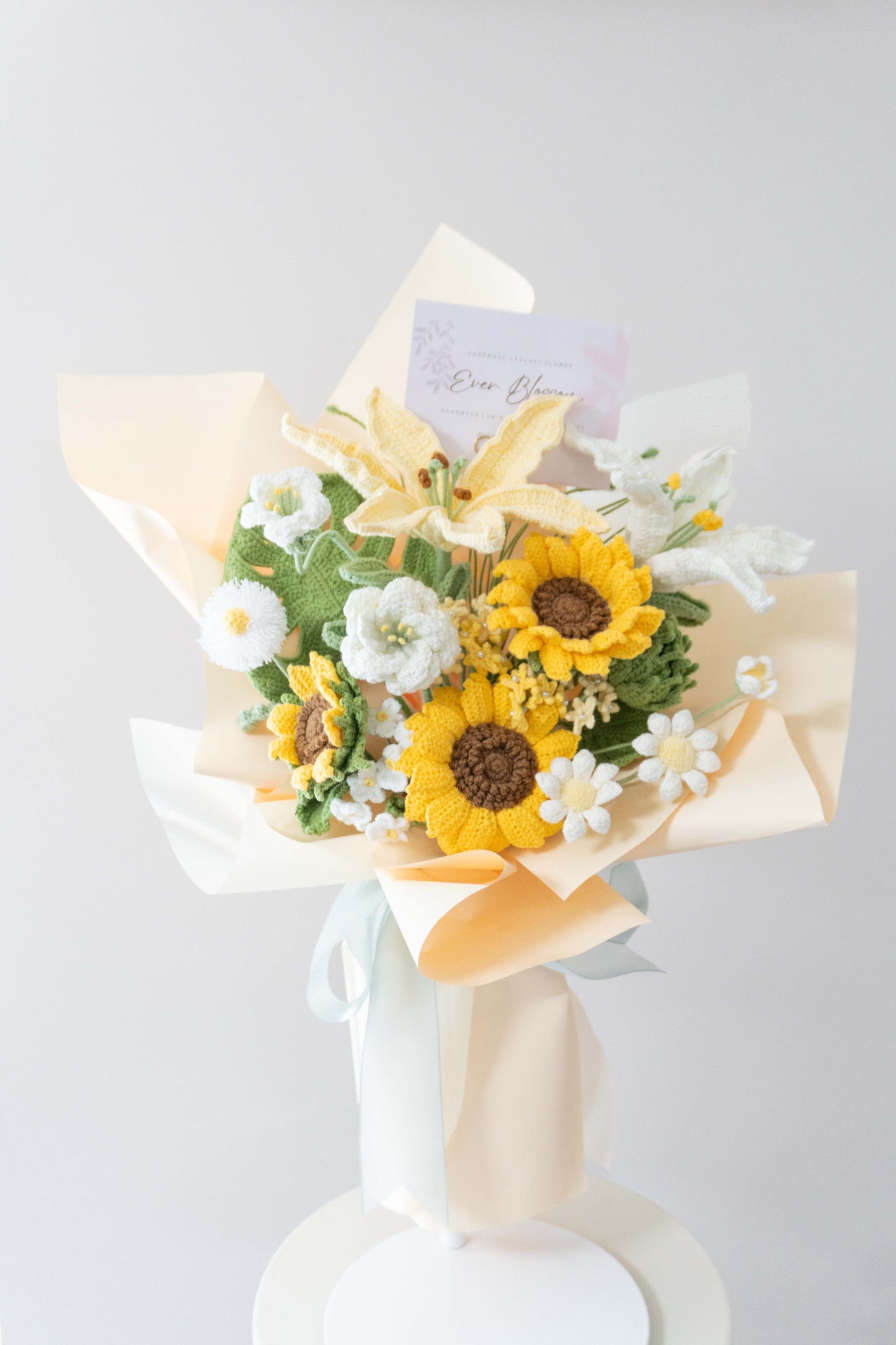 這是一束由香港花店Ever Blossom精心製作的永生花束，結合向日葵象徵追求幸福與百合代表優雅高貴，搭配白色雛菊與綠葉，包裹在淡雅薄紙和藍色緞帶中。花束設計精緻，展現溫暖與高雅氣息，適合用作生日、紀念日或送禮選擇。永生花採用高品質材料編織，保存時間長，可作為家居裝飾或表達心意的完美禮物，體現香港花店的專業工藝與創意。